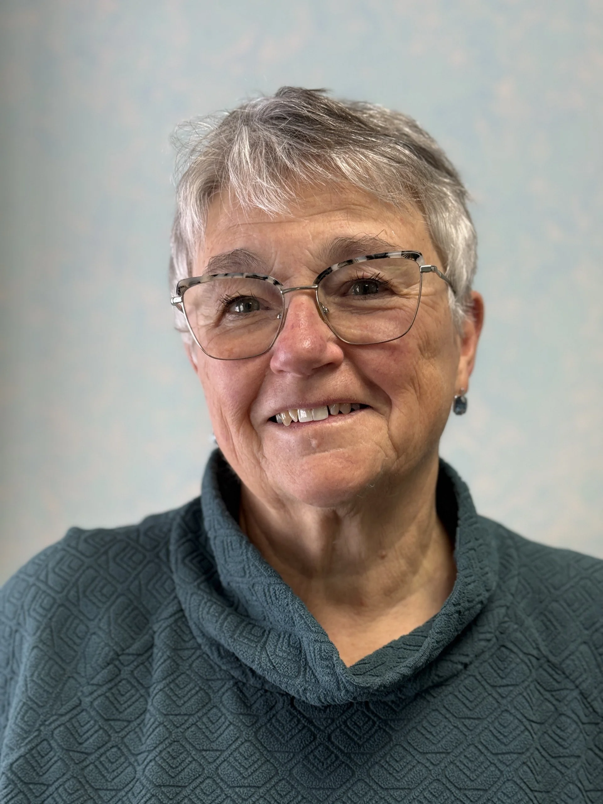 A smiling elderly woman with short gray hair, glasses, and a dark patterned sweater, standing against a light-colored background.