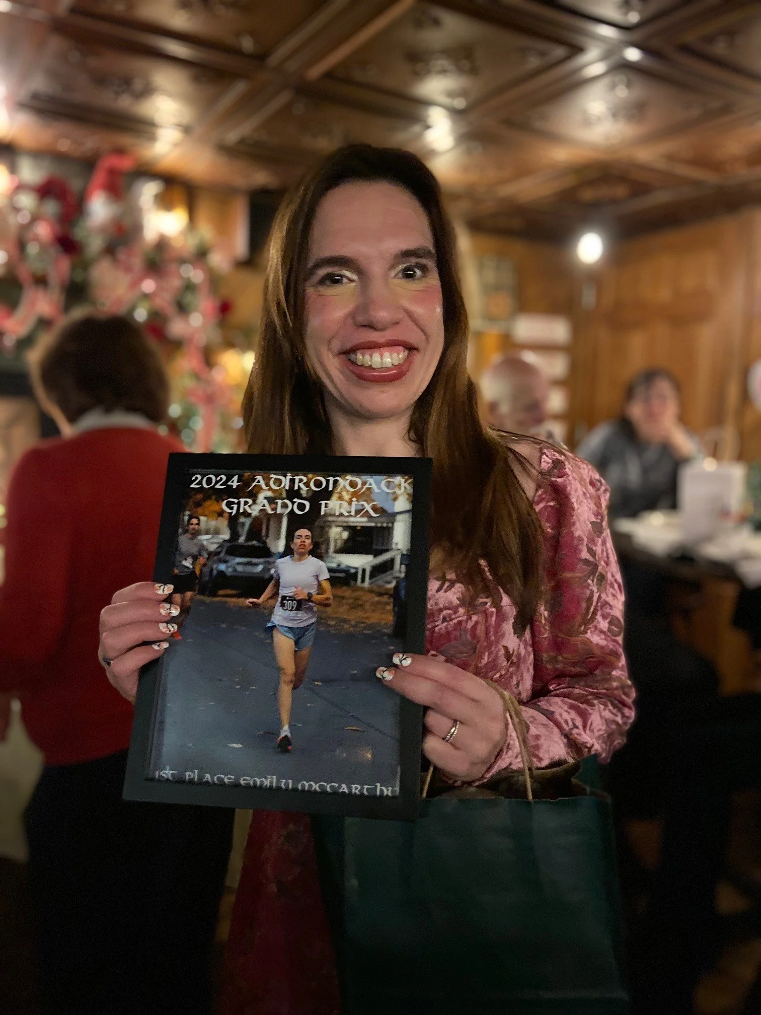 A woman with long brown hair smiling and holding a photo of an award in front of a Christmas tree inside a wooden-paneled room.