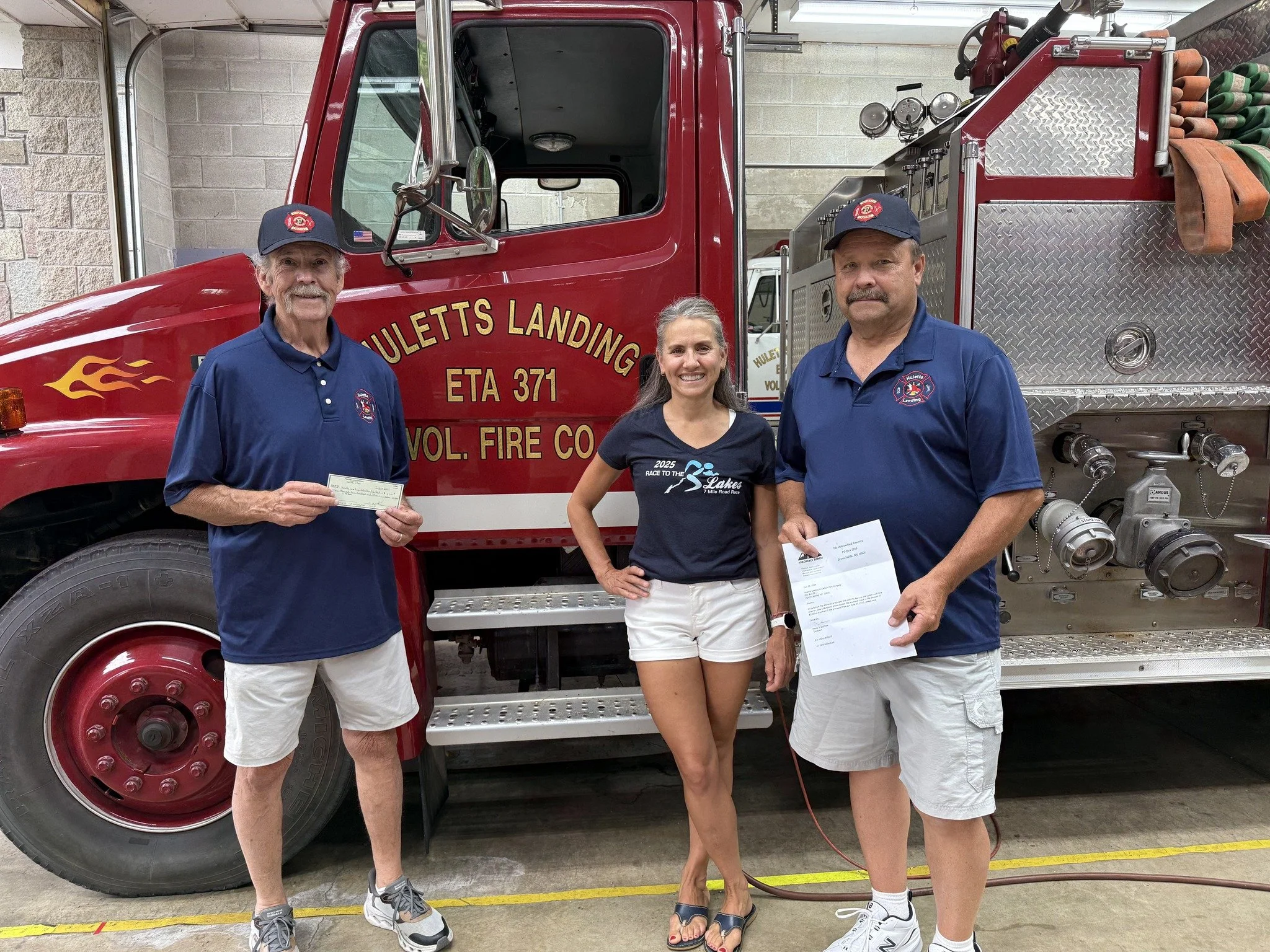 Three people standing in front of a red fire truck, with two men wearing navy blue shirts and caps, and a woman in a black t-shirt and white shorts, all smiling. The man on the left holds a check, the woman has her hand on her hip, and the man on the right holds papers. The fire truck is labeled "Hulett Landing Volunteer Fire Co."