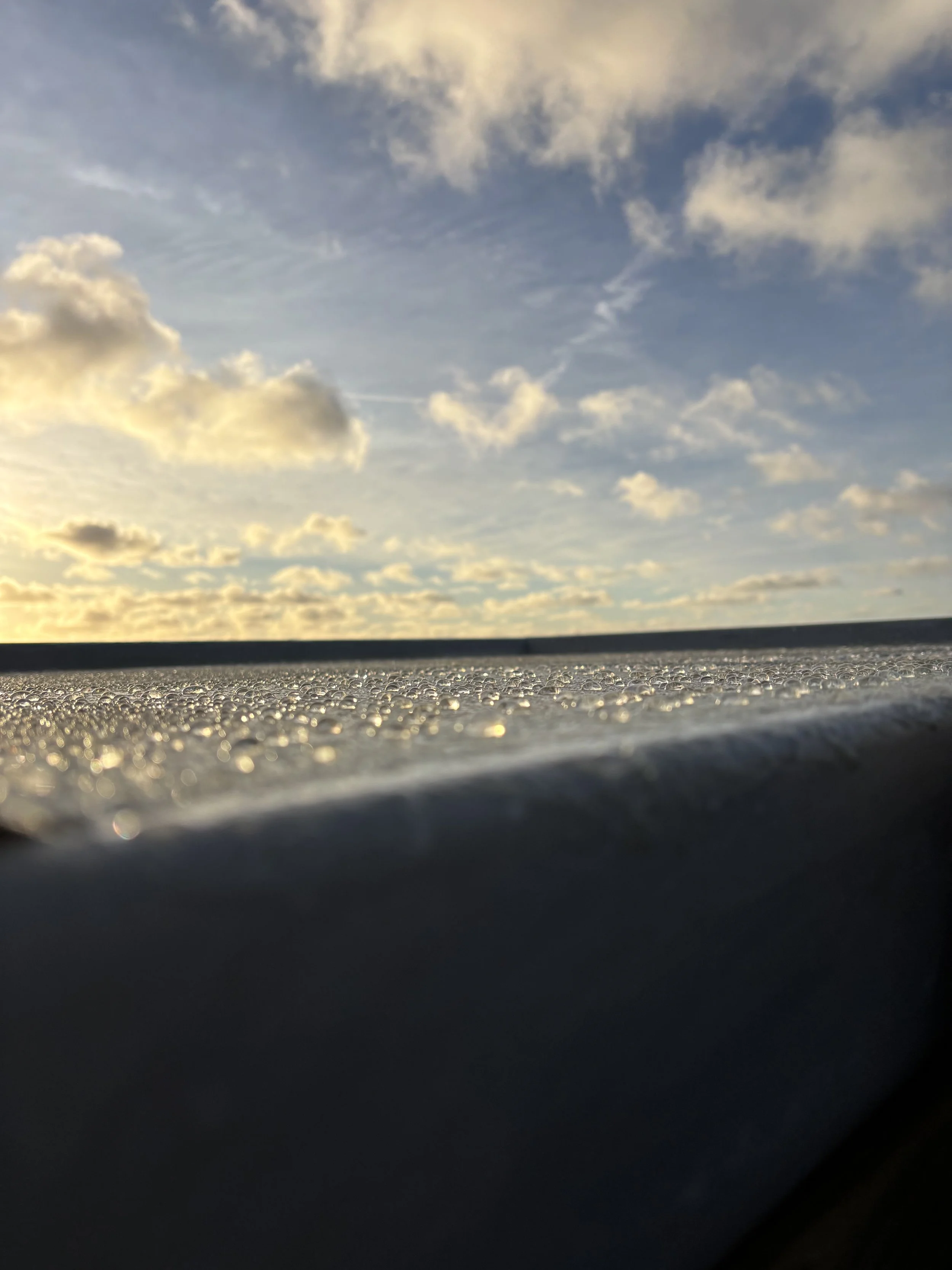 Close-up view of a textured surface with water droplets, with a sky filled with clouds and a setting sun in the background.