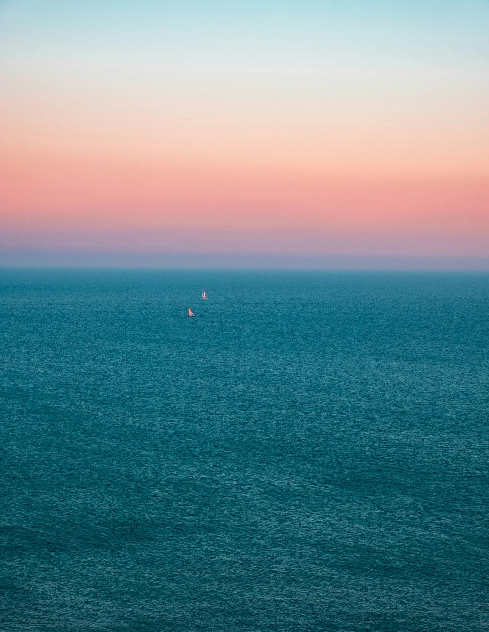 A calm ocean with two sailboats in the distance under a colorful sky with pink, purple, and faded blue hues.