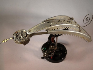A mechanical or robotic insect made from metallic and electronic parts, resembling a combination of a dragonfly and beetle with a curved, segmented body and green eyes.