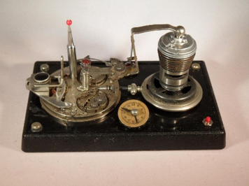 A vintage electrical device with metal components, gears, and a small circular gauge mounted on a black base.