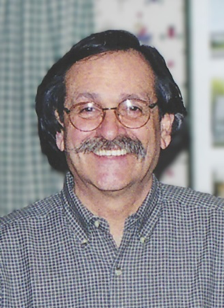 A middle-aged man with dark, wavy hair and a mustache, wearing glasses and a checkered shirt, smiling indoors with a blurred background.