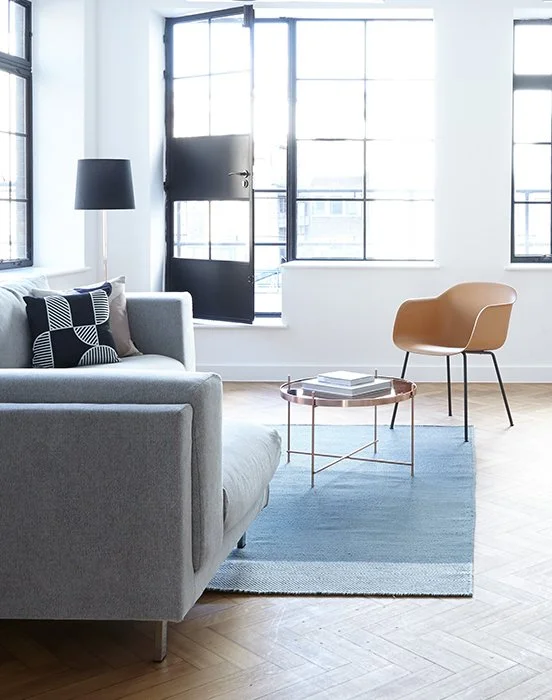 Modern living room with gray sofa, black and white patterned pillow, black floor lamp, beige chair, glass-top coffee table with books, blue rug, large windows, and black door, in a bright, minimalist style.
