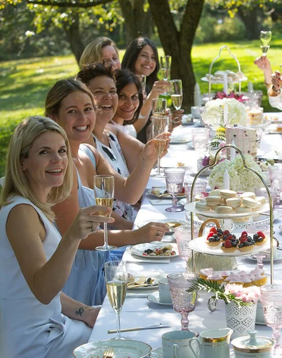 Women celebrating outdoors at a garden tea party with champagne and desserts on a long table.