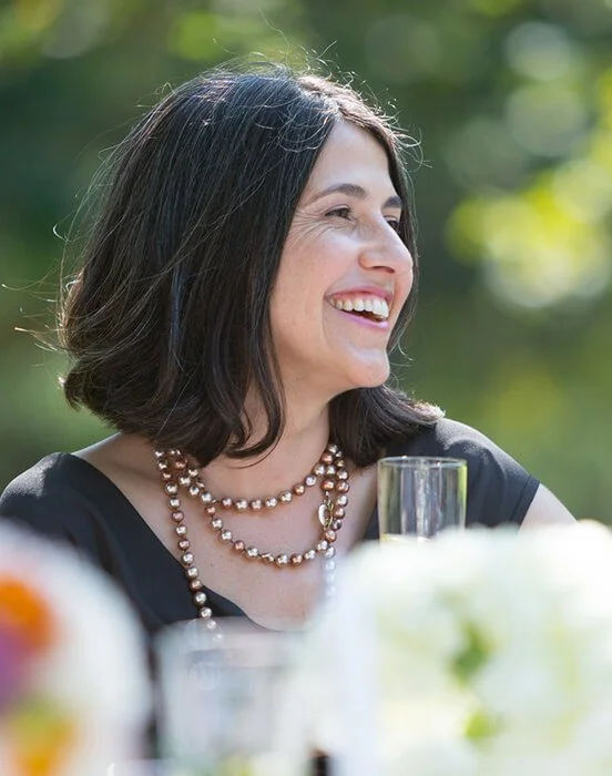 A woman with dark hair, smiling and wearing pearl necklaces, at an outdoor event with greenery in the background.