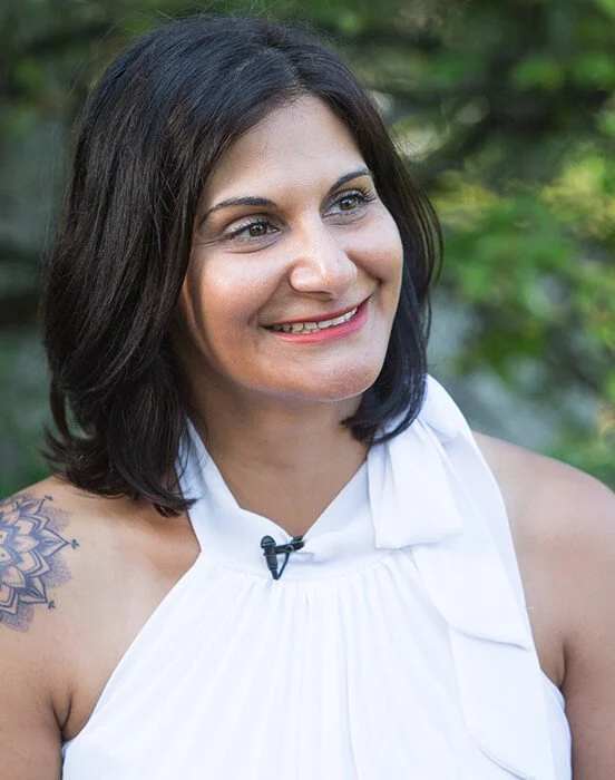 A woman with shoulder-length dark hair smiling outdoors, wearing a white sleeveless top with a bow detail and a visible tattoo on her left shoulder, with greenery in the background.