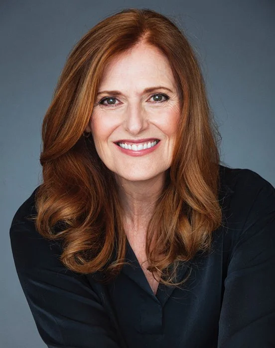 A woman with long red hair smiling, wearing a black top, against a gray background.
