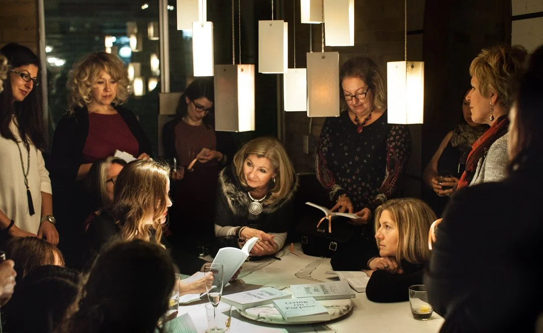 A group of women gathered around a woman sitting at a table, engaged in conversation, in a warmly lit indoor setting.