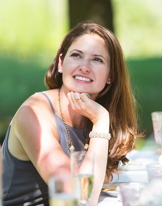 A woman with long brown hair, smiling and looking to the side, sitting outdoors at a table with drinks.