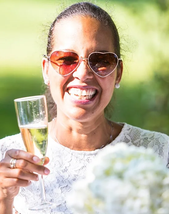 Woman smiling, wearing heart-shaped sunglasses, holding a glass of champagne, outdoors with greenery.