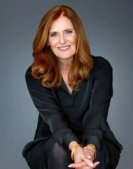 A woman with long red hair wearing a black dress, smiling, sitting against a gray background.