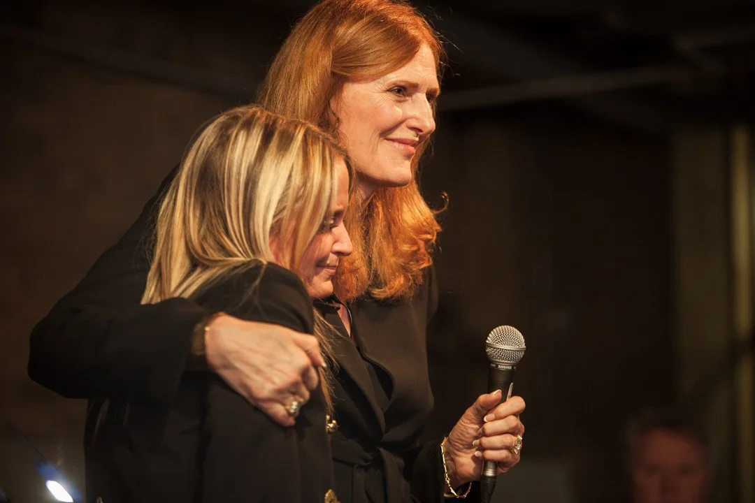Two women hugging on stage, one holding a microphone, with warm lighting in the background.