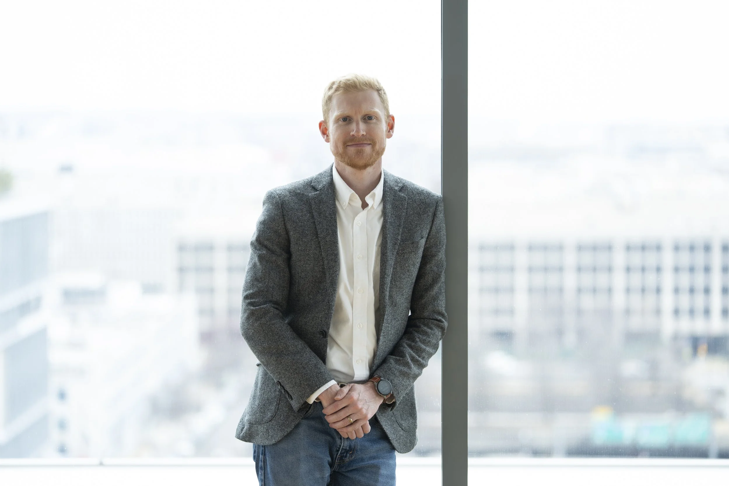 Nick Coe, a man with red hair and a beard wearing a gray blazer, white shirt, and jeans, standing indoors next to a large window with a cityscape in the background.