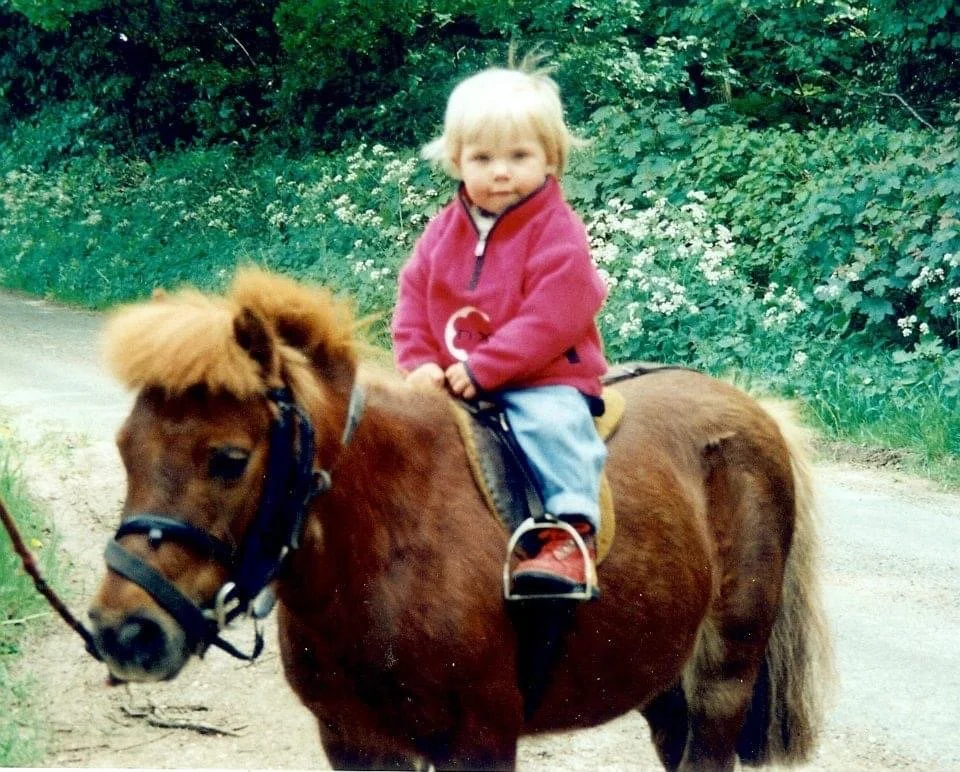 Young child with blonde hair riding a small brown pony on a dirt path, surrounded by greenery and white flowers.
