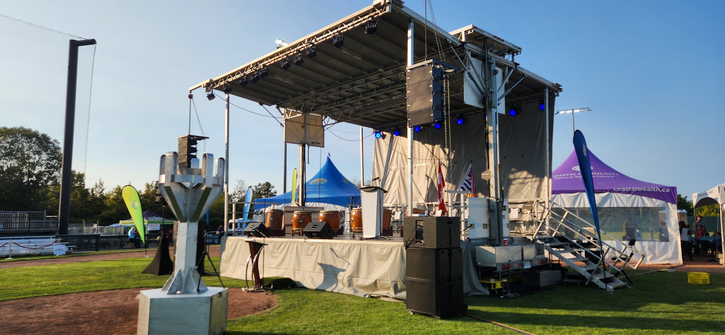 Empty outdoor stage set up for an event with drums, audio equipment, nearby tents, flags, and a scenic outdoor park with grass and trees.
