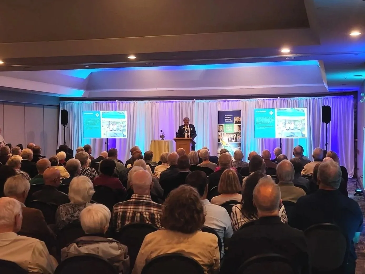 A speaker giving a presentation to a large audience seated in a conference room with two screens displaying slides.