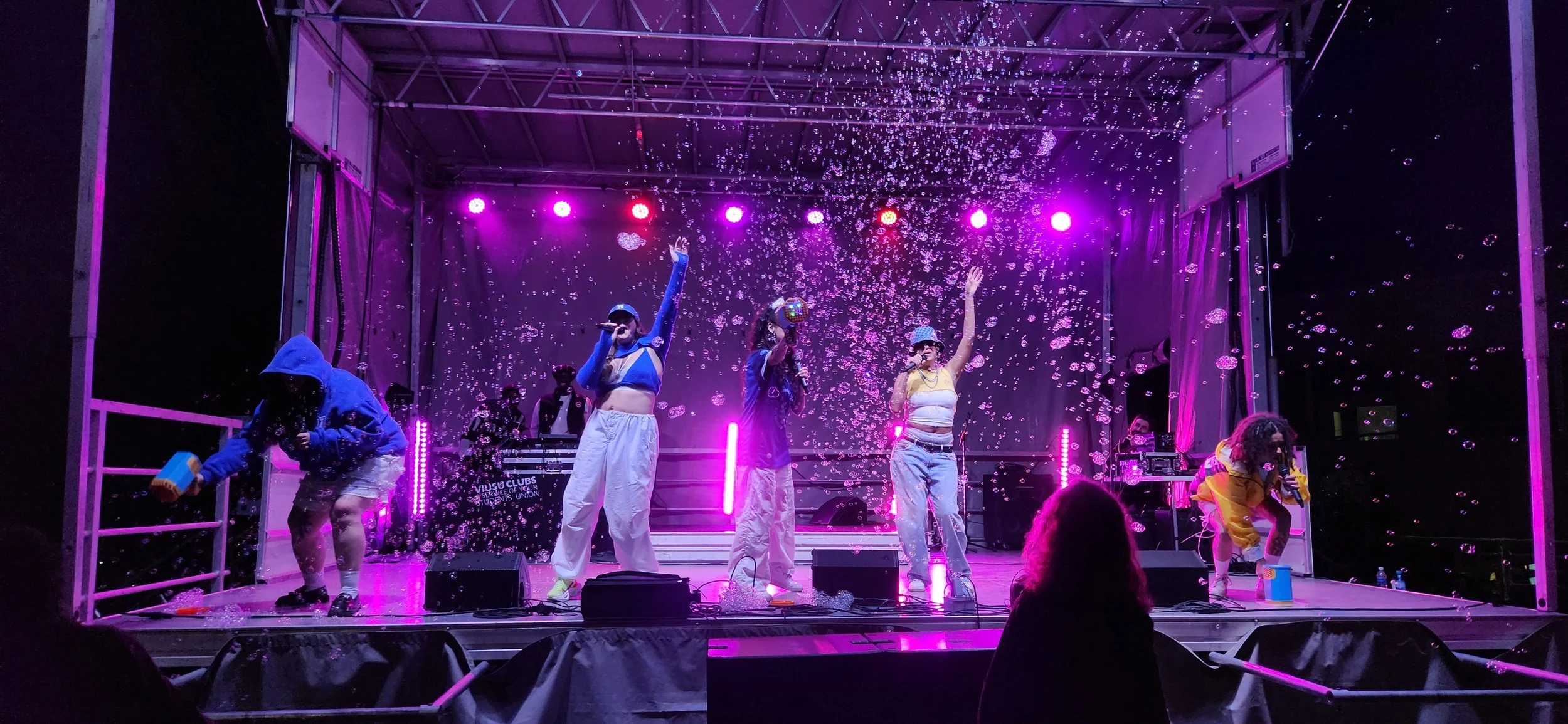 A live concert stage with five performers under purple and pink lighting, surrounded by falling bubbles, with DJ equipment and musicians in the background.