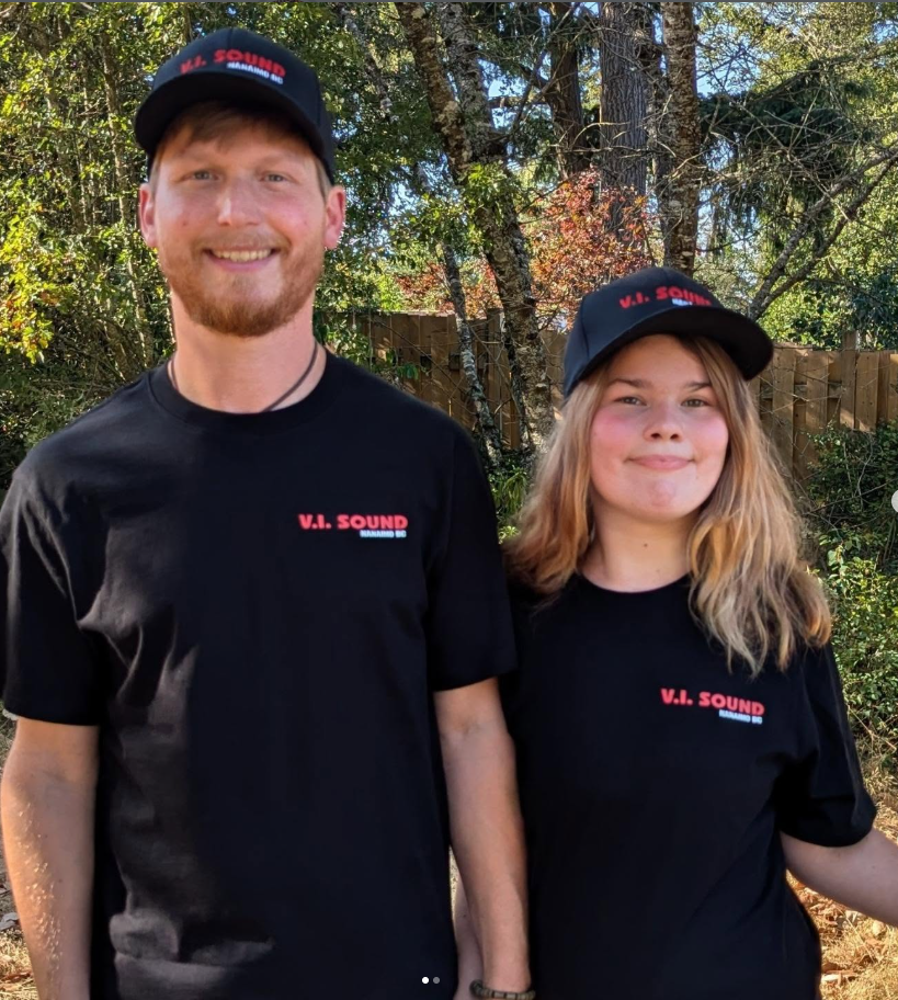 A young man and woman smiling outdoors wearing black V.I. Sound T-shirts and caps, standing in front of trees and a wooden fence.