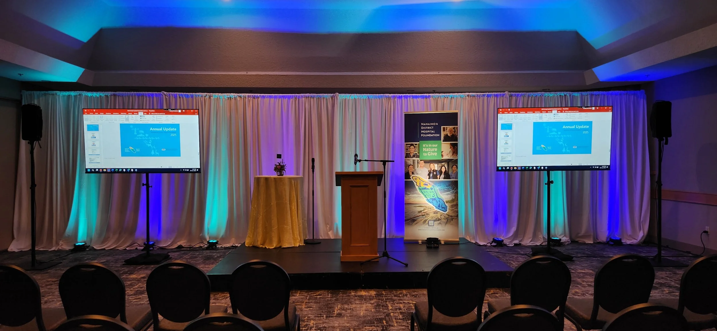 Empty stage with a wooden podium, two large screens displaying a presentation titled 'Annual Update 2023,' a microphone, a small round table with a bouquet, and a colorful banner with a map and images of diverse people, set against a white curtain backdrop with blue and green lighting.