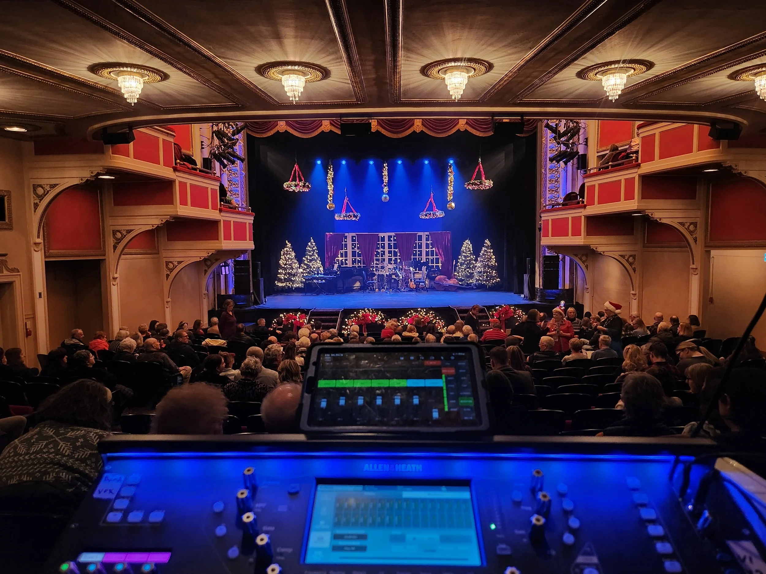 The image shows a theater decorated for Christmas with lit Christmas trees, hanging decorations, and wrapped presents. The stage is set with purple curtains and musical instruments. Audience members are seated, and there is audio equipment in the foreground.