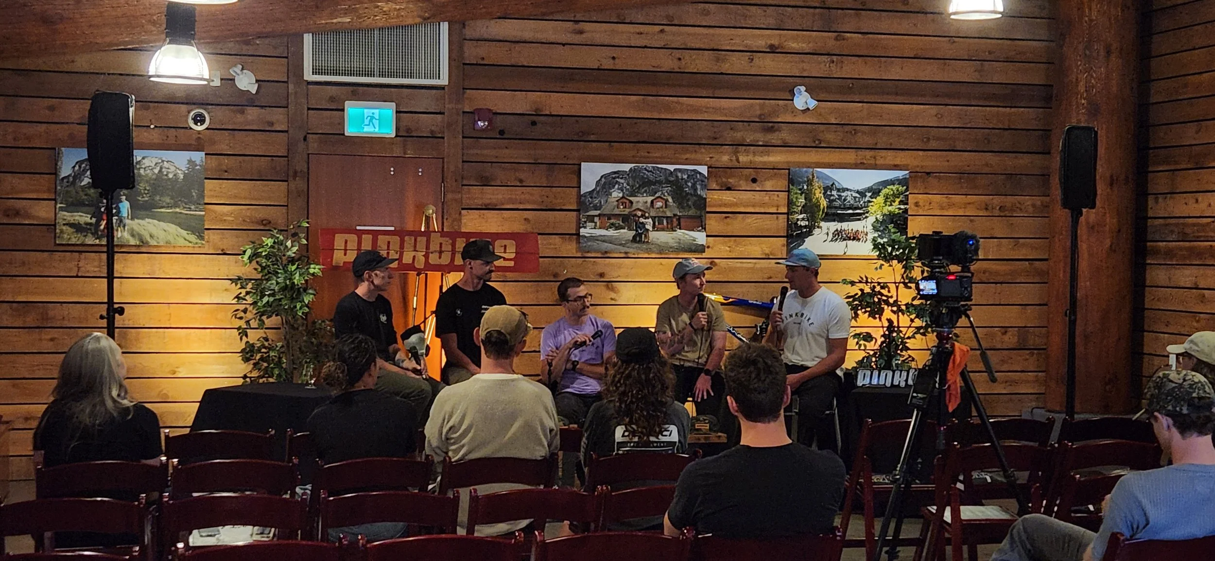 Indoor event with panel of five people on stage, audience seated in chairs, wooden walls, artwork, and filming equipment.
