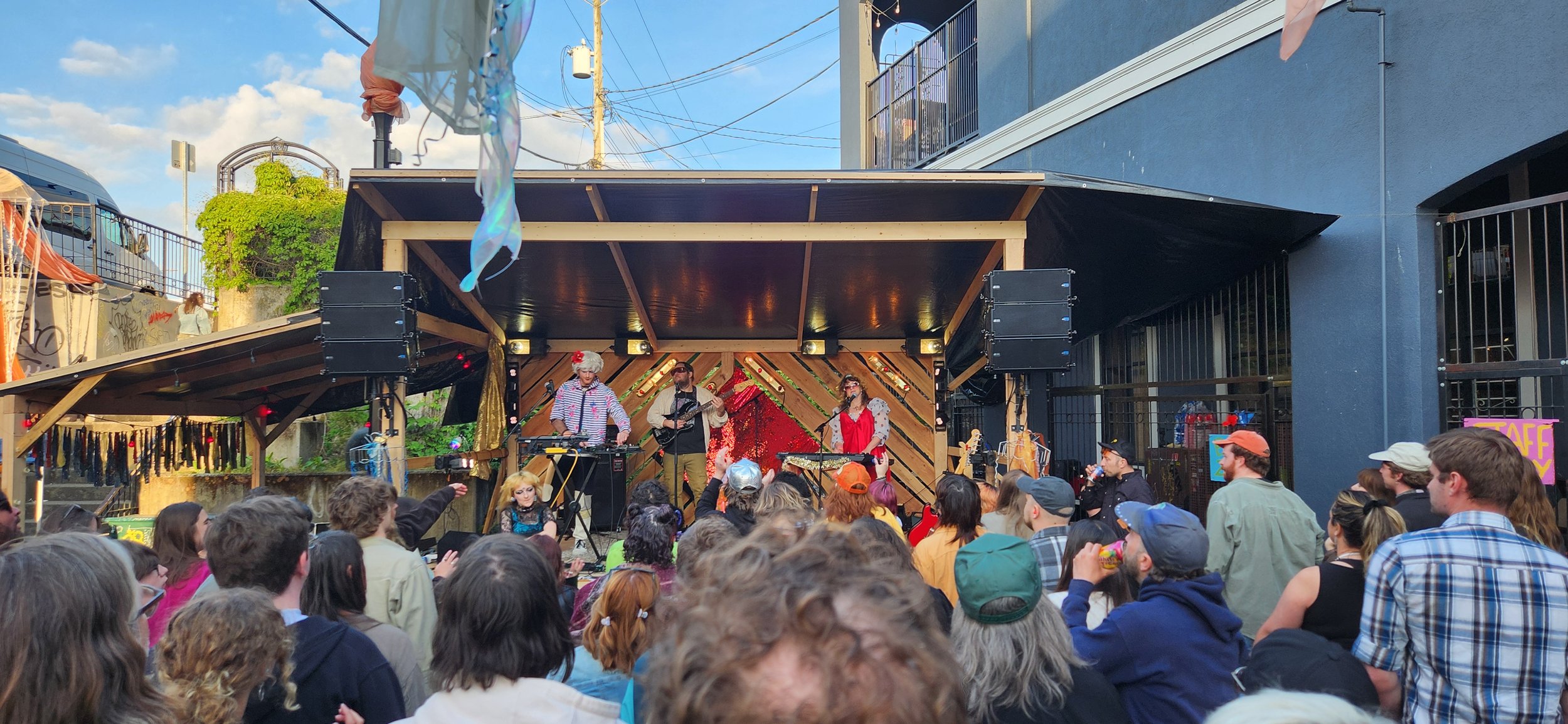 Outdoor live music concert with a diverse crowd of people watching a band perform on a small stage decorated with colorful curtains and red sequined fabric. The band members wear vintage costumes, and the audience is standing, some with hats and jackets, enjoying the event.