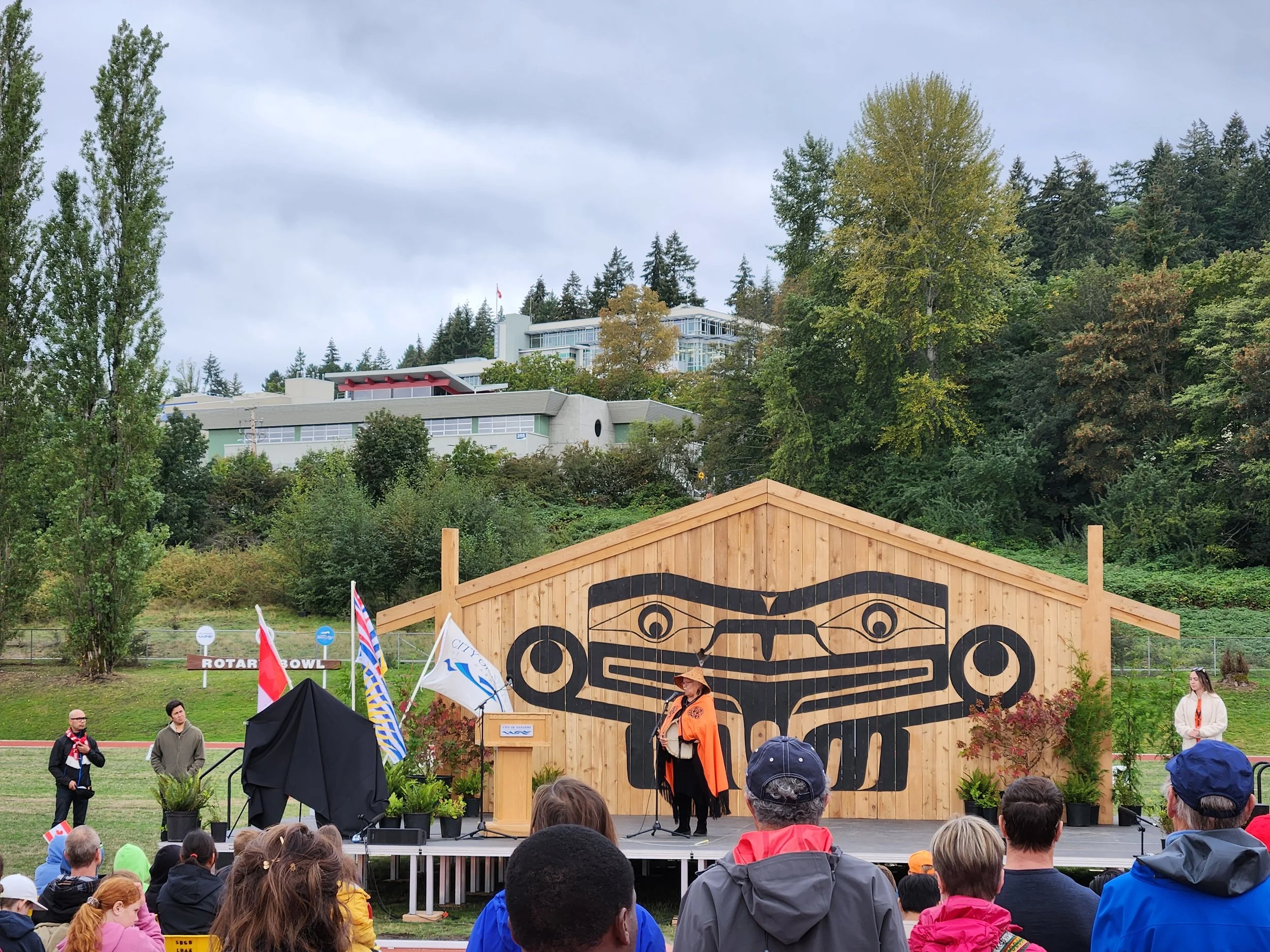 Outdoor gathering with a stage in front of a wooden totem pole art installation, people in the audience, and a person speaking on stage. Flags and potted plants are visible on stage, with trees and buildings in the background.