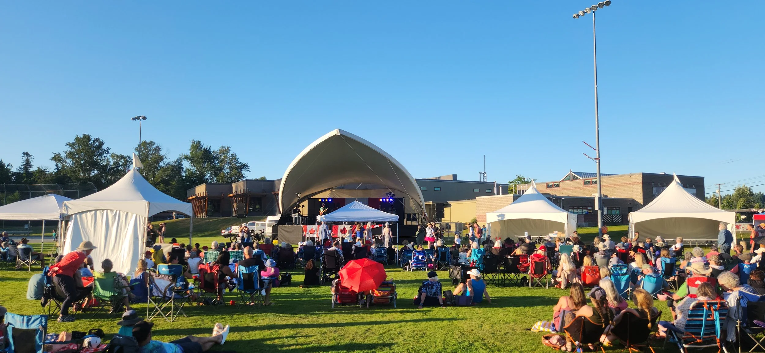 Outdoor concert on a sunny day with a large crowd seated on grass in front of a stage with performers. Several white tents are set up around the area, and a red umbrella provides shade for some attendees. The background includes trees and modern buildings.