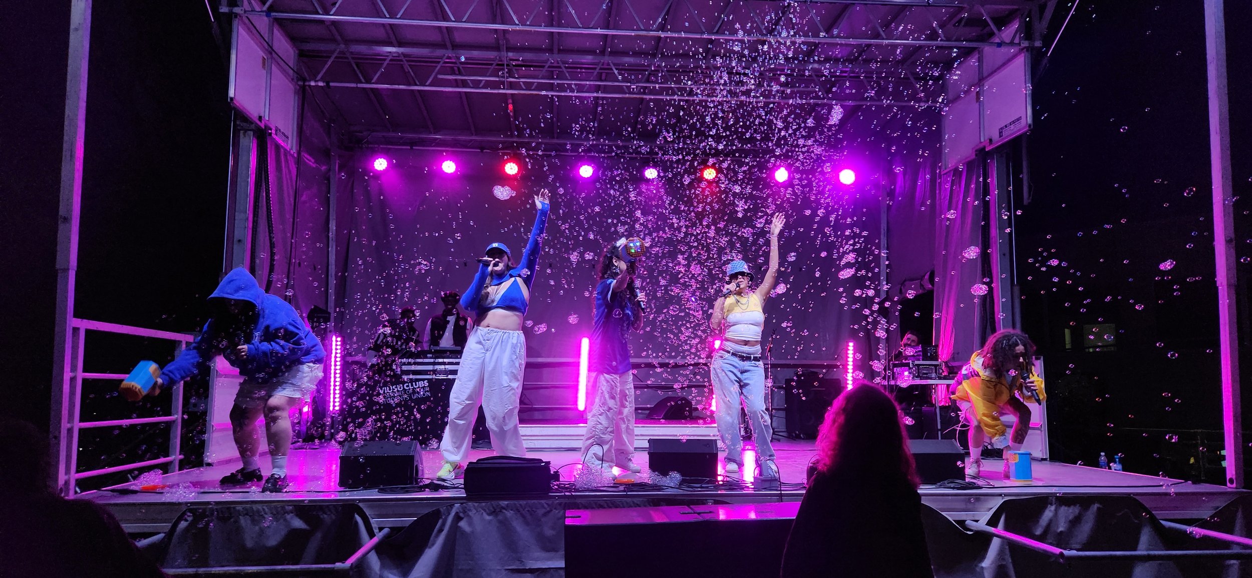 A group of performers on stage at night, with purple and pink lighting, wearing colorful casual clothing, surrounded by bubbles and sound equipment, engaging in a lively performance.