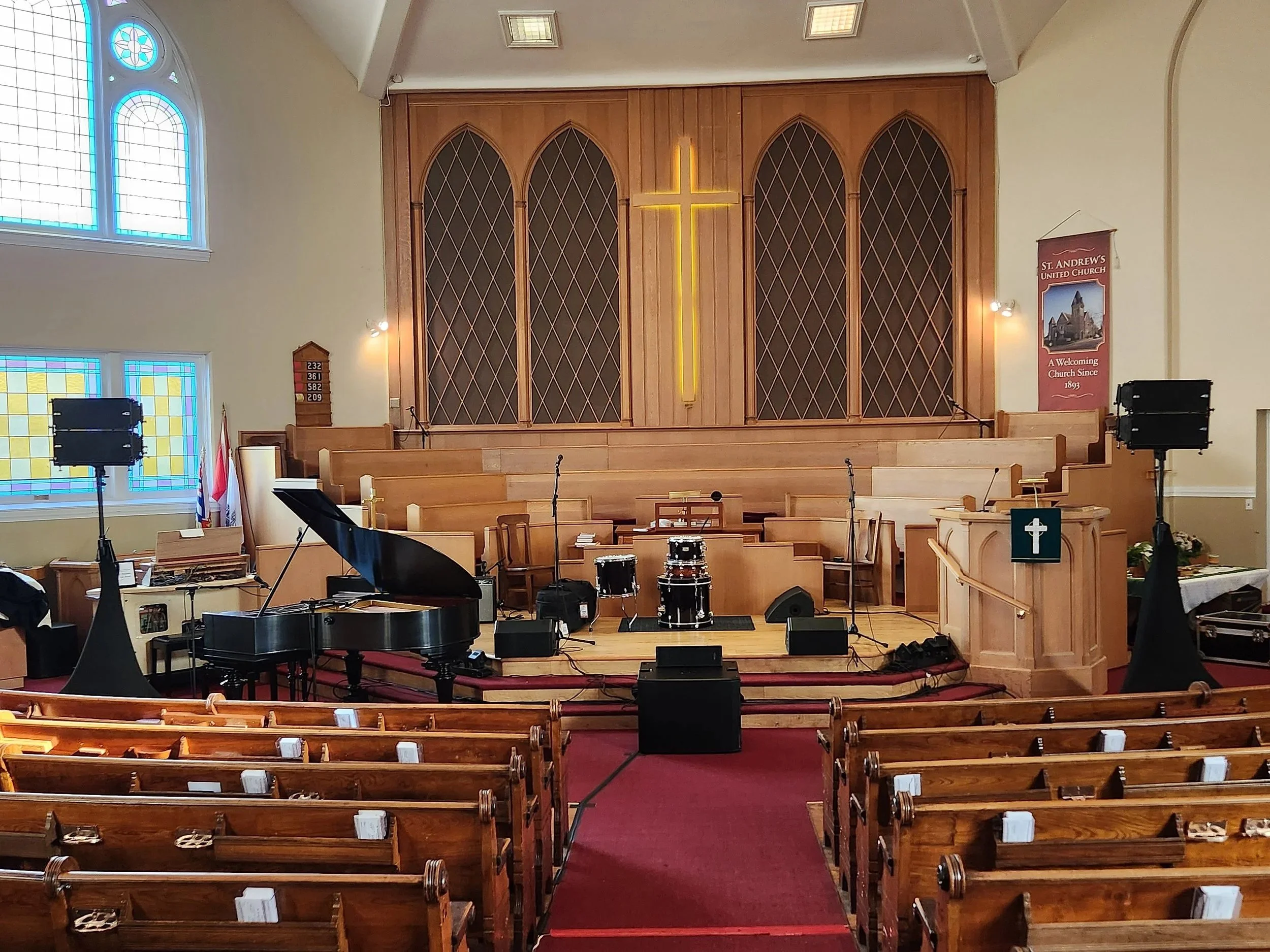 The interior of a church sanctuary, featuring a stage with musical instruments including a piano, drums, microphones, and speakers. A large wooden cross is illuminated and positioned at the center on the back wall. There are stained glass windows on the left side and banners on the right, with wooden pews in the foreground.