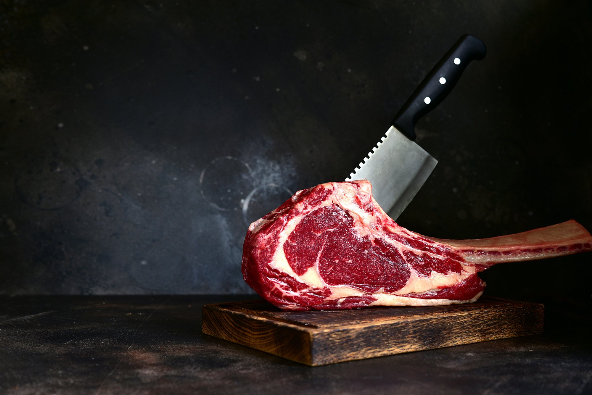 A raw, marbled beef steak on a wooden cutting board with a black knife set on a dark background - Quality meats at Dickenson's of Fulham