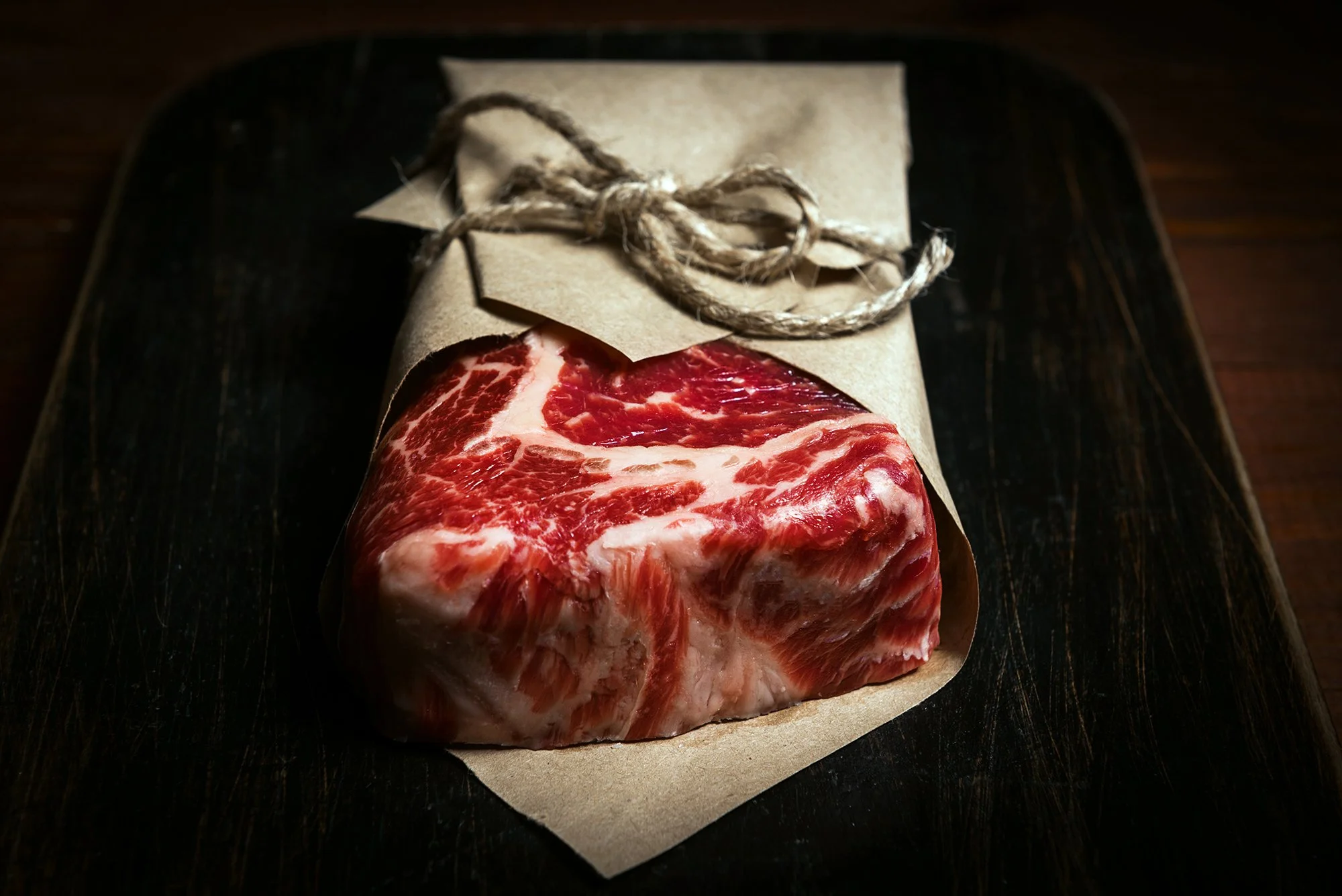 A raw cut of marbled beef resting on brown parchment paper with a rustic twine tied around it, placed on a dark wooden surface - Quality meats at Dickenson's of Fulham