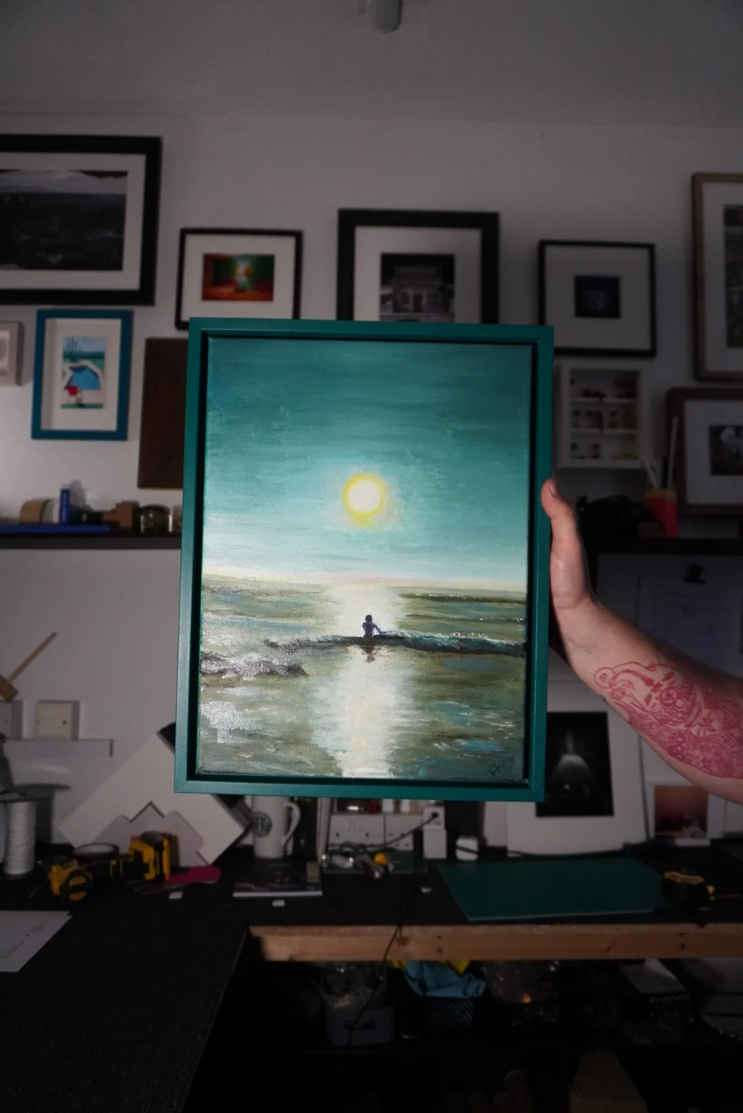 A person holding up a painting of a seascape with the sun reflecting on the water, with a background of framed pictures on a wall.