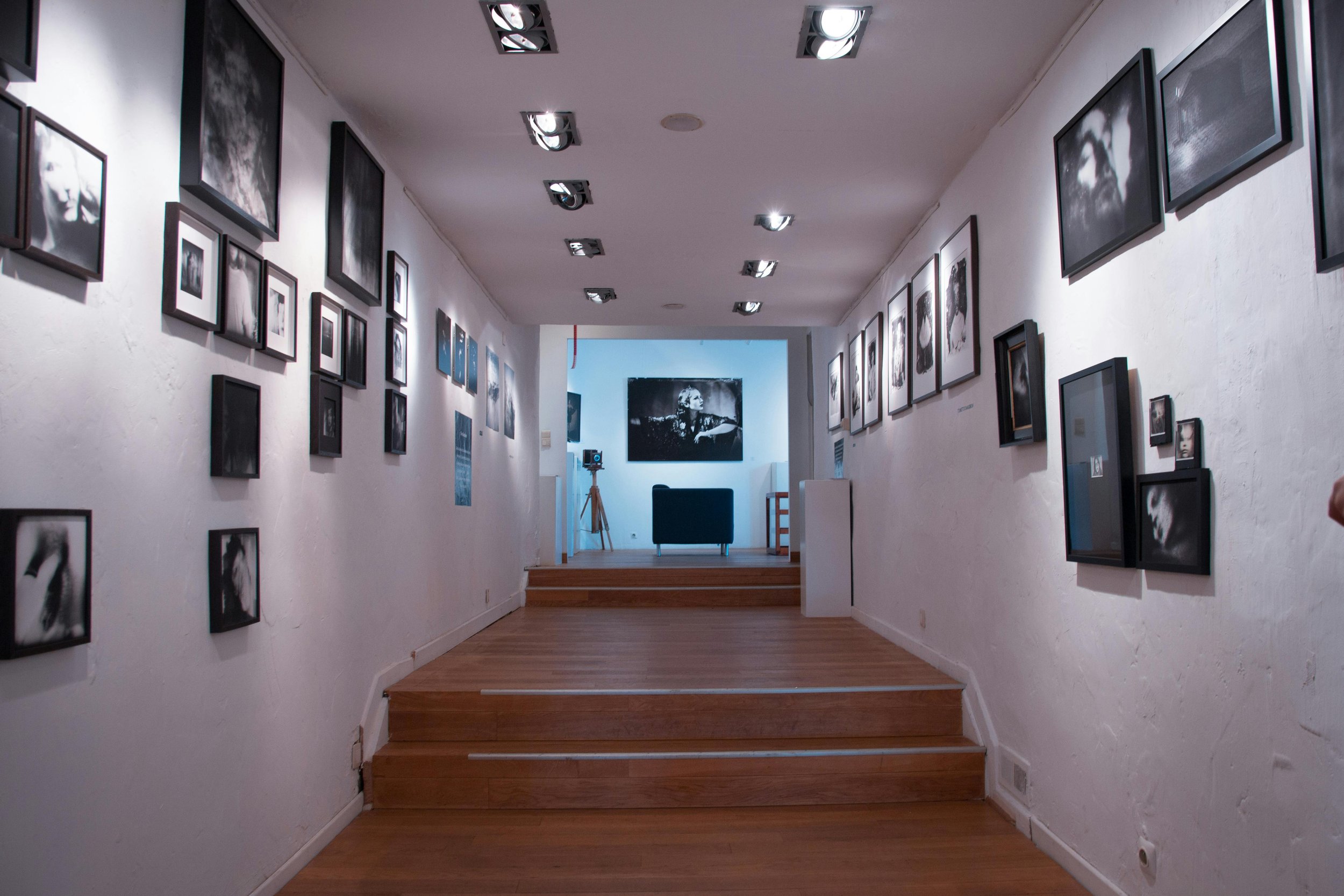 An art gallery hallway with black-and-white photographs on the white walls, wooden floor, and ceiling lights. There are steps leading up to a small platform with more photographs and art pieces at the far end.