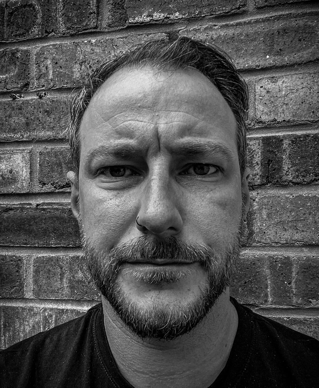 Black and white close-up portrait of a man with a beard and mustache, wearing a nose ring, standing in front of a brick wall.
