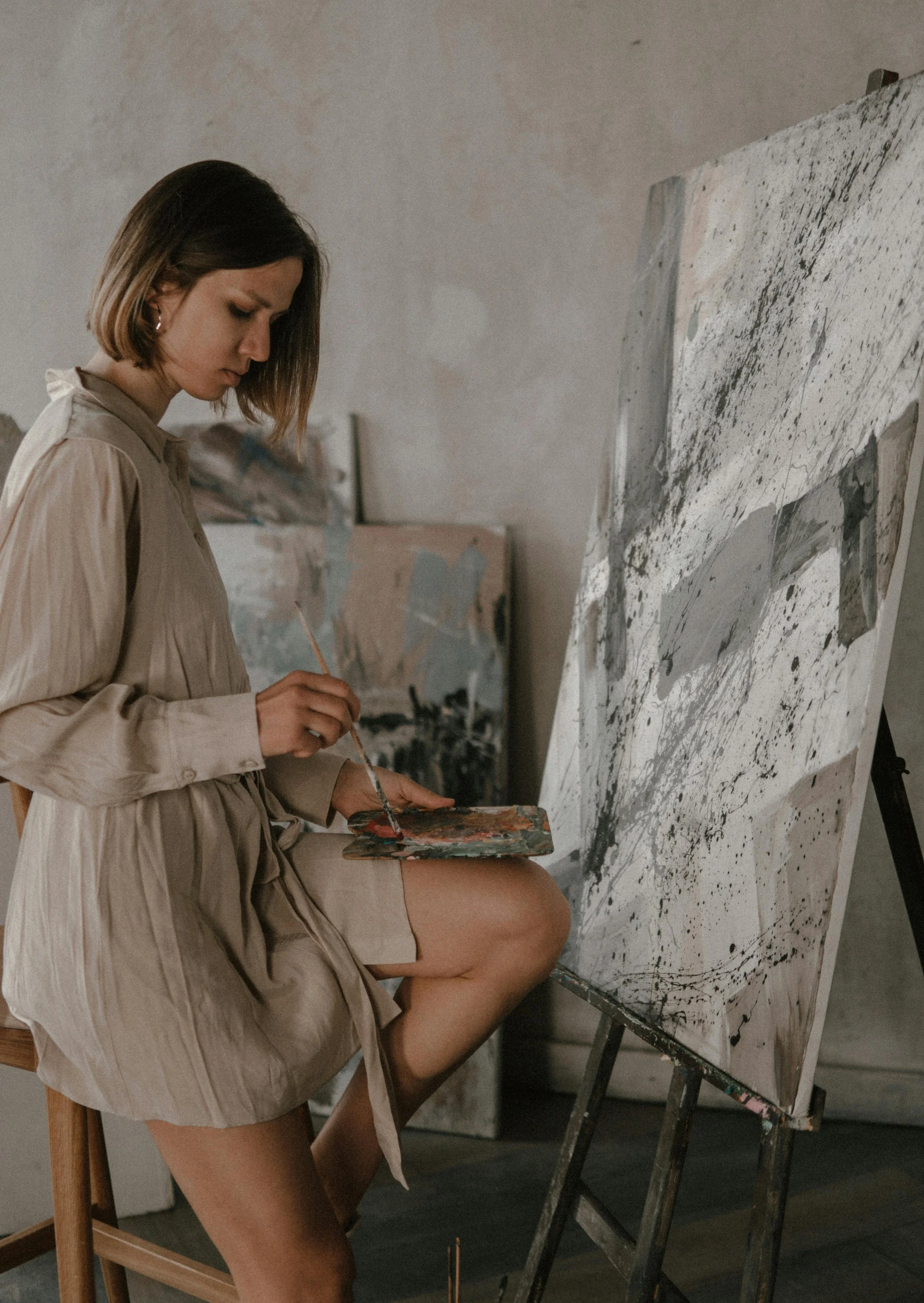 A woman is sitting on a stool, working on a large abstract painting with black, white, and gray splashes and lines on an easel.