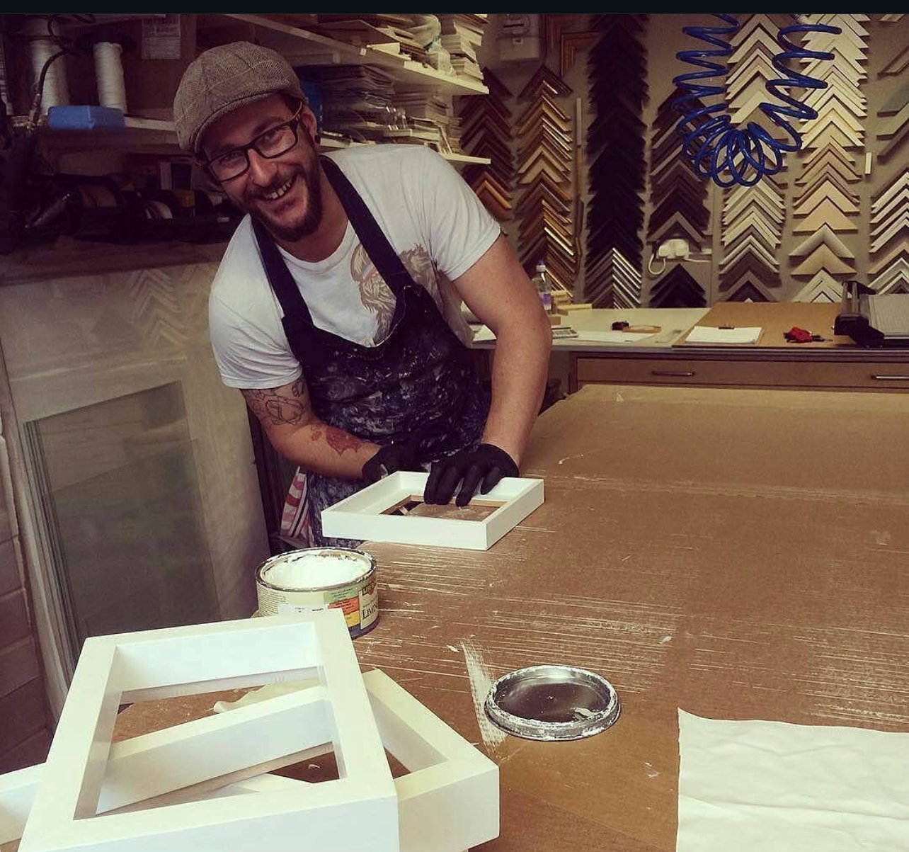 A man with glasses, a cap, and tattoos, smiling while working on a picture frame in a woodworking shop, surrounded by tools, paint, and frame samples.