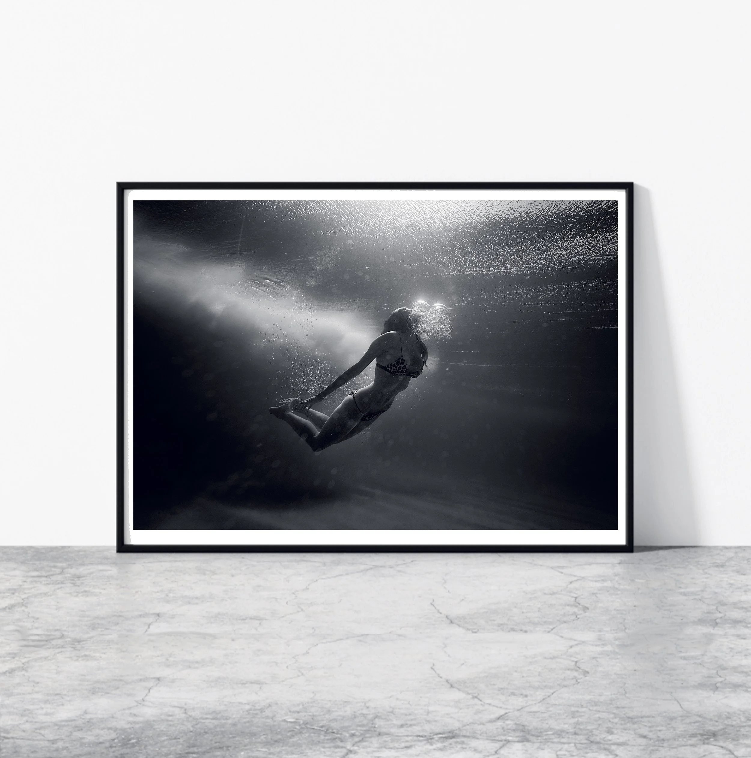 Black and white photograph of a woman swimming underwater, wearing a bikini, looking upward, with light rays shining through the water.