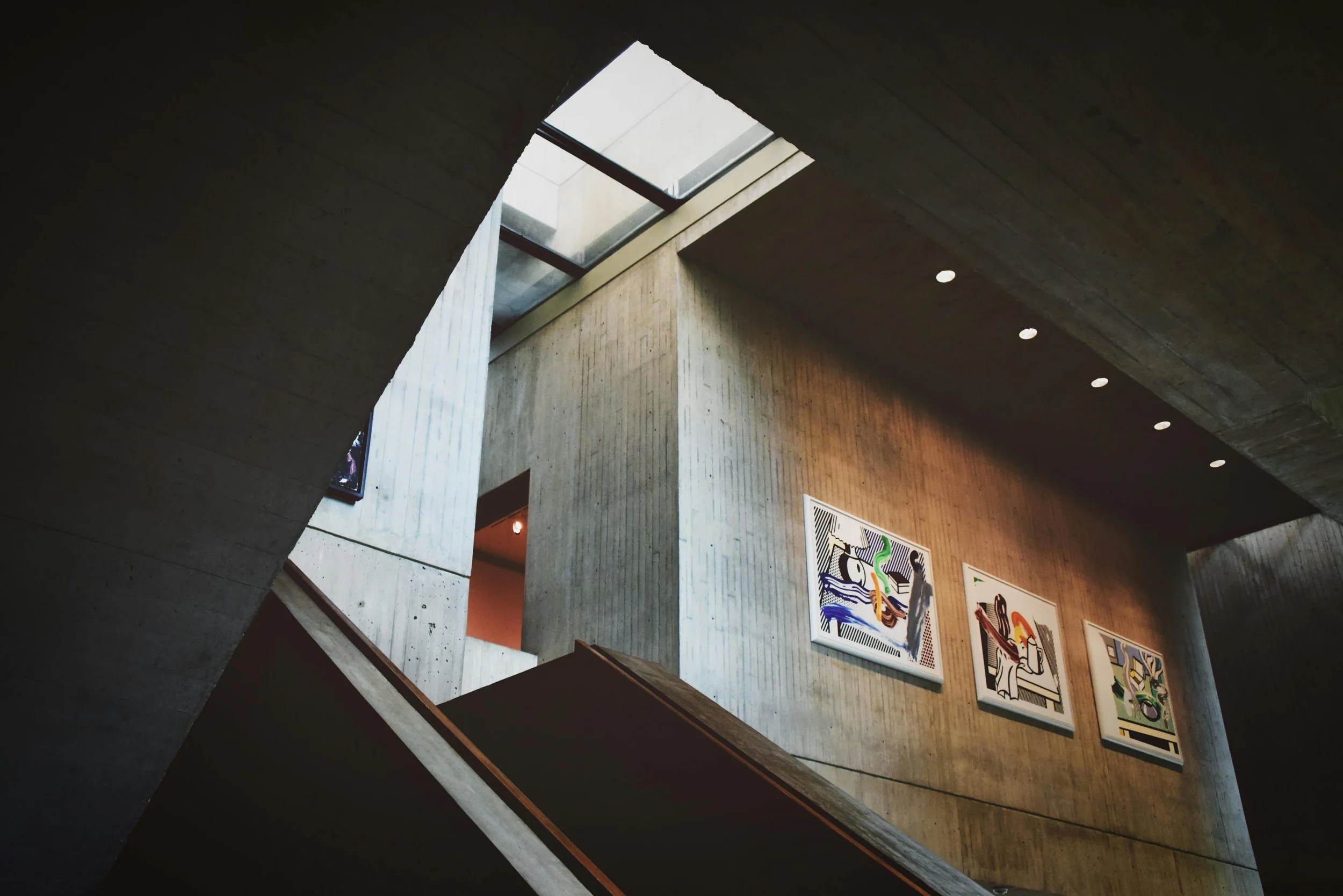 Interior of a modern art museum with concrete walls, colorful abstract paintings on display, skylight above, and an angled staircase.