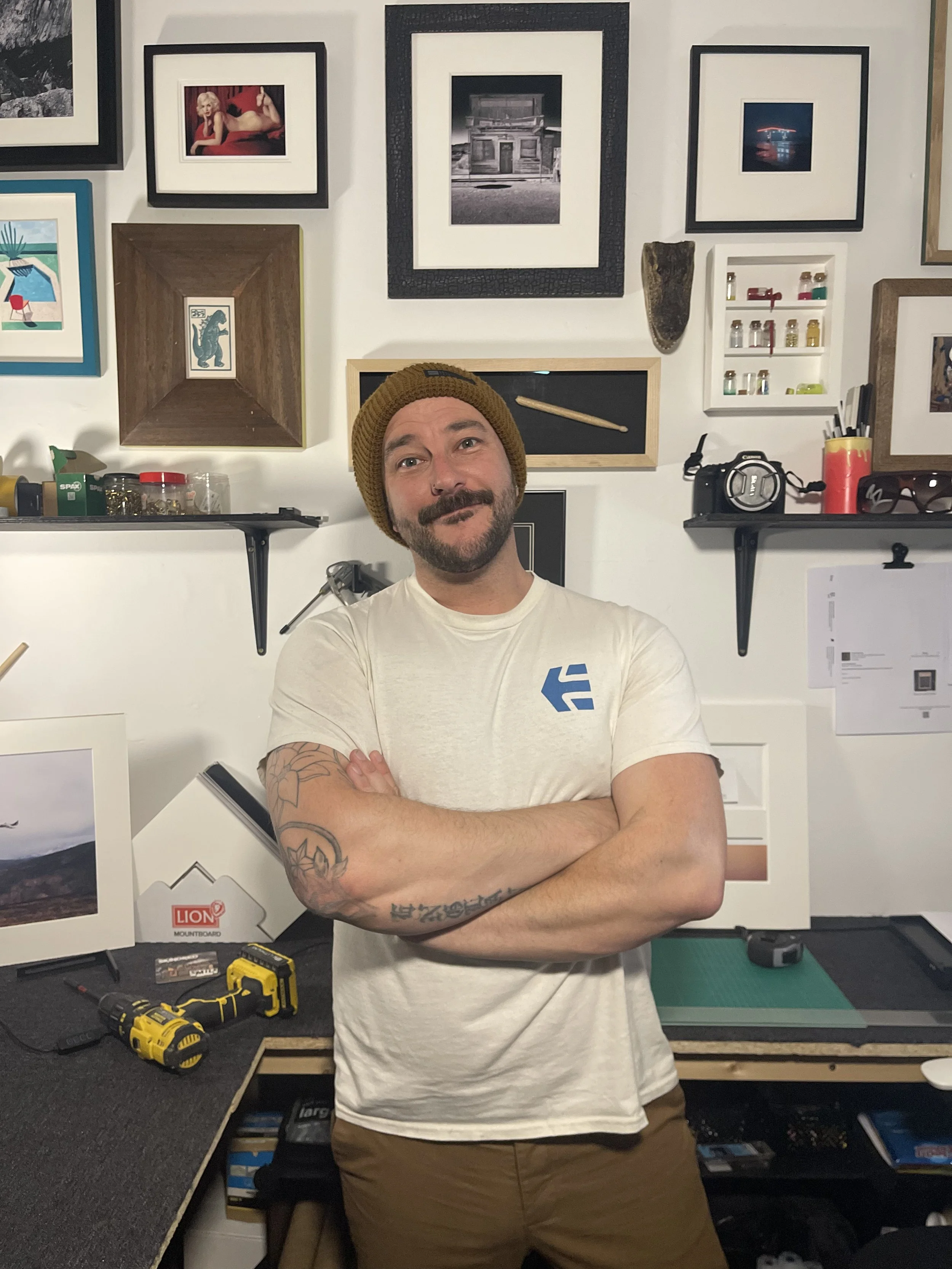 A man with a beard and mustache wearing a brown beanie and a light-colored t-shirt, standing with his arms crossed in a room with framed photos and artwork on the wall behind him. The workspace has tools, supplies, and framed pictures.