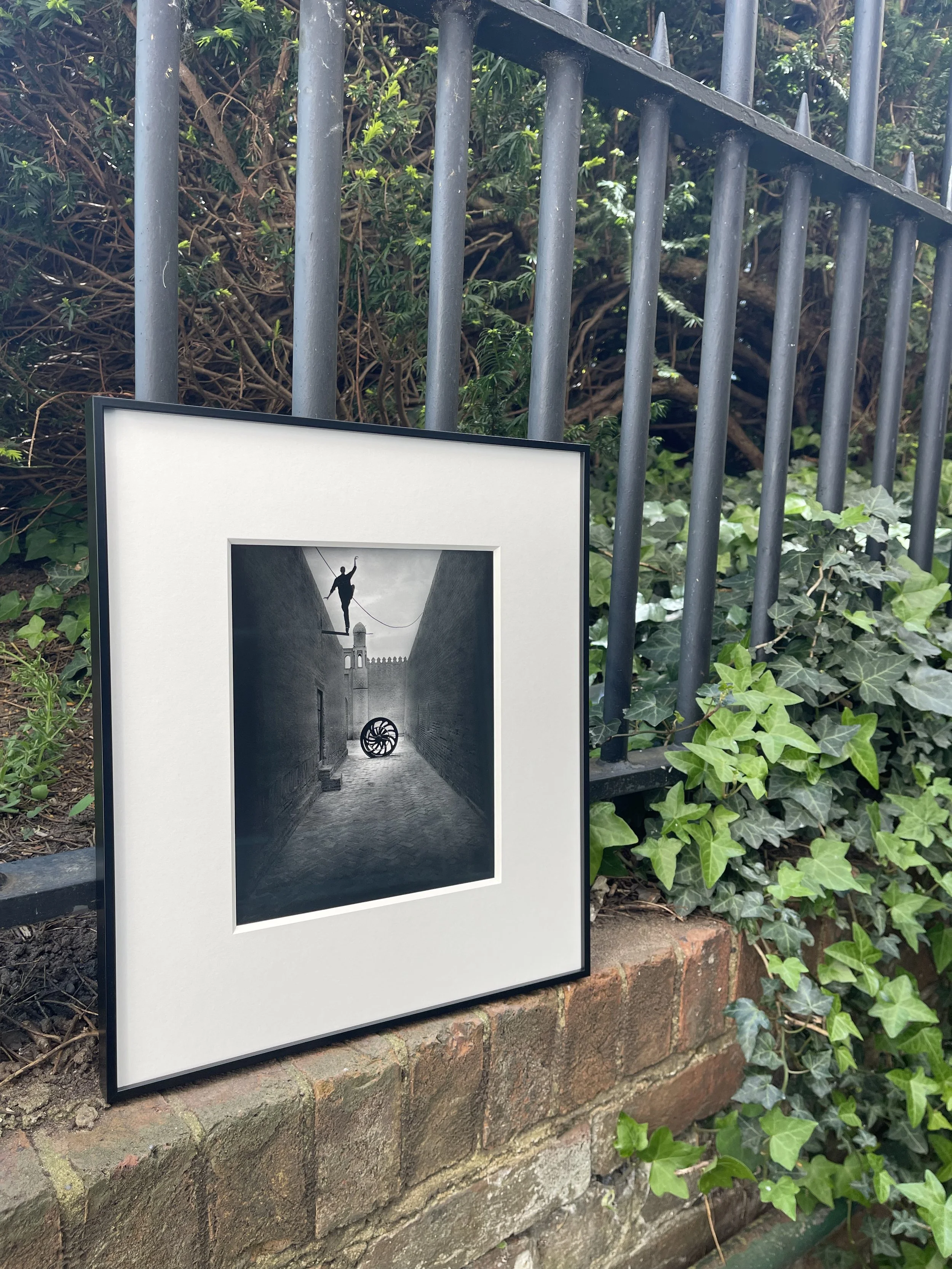 Framed black and white surreal photo of a narrow alleyway, with a person balancing on a tightrope between two walls, and a large spinning wheel on the ground at the end of the alley.
