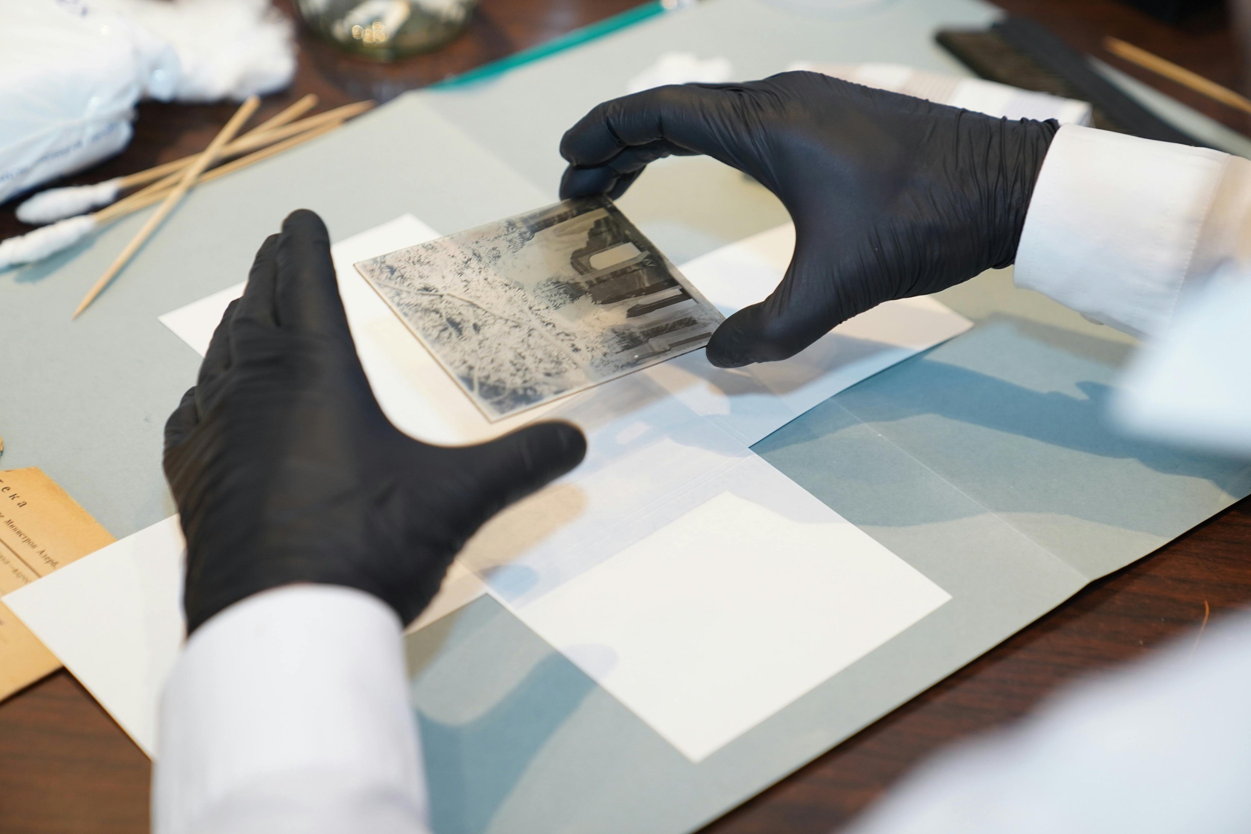 A person wearing black gloves and a white lab coat handling a glass plate with an image of a forest landscape, possibly for preservation or analysis, on a table with various tools and materials around.