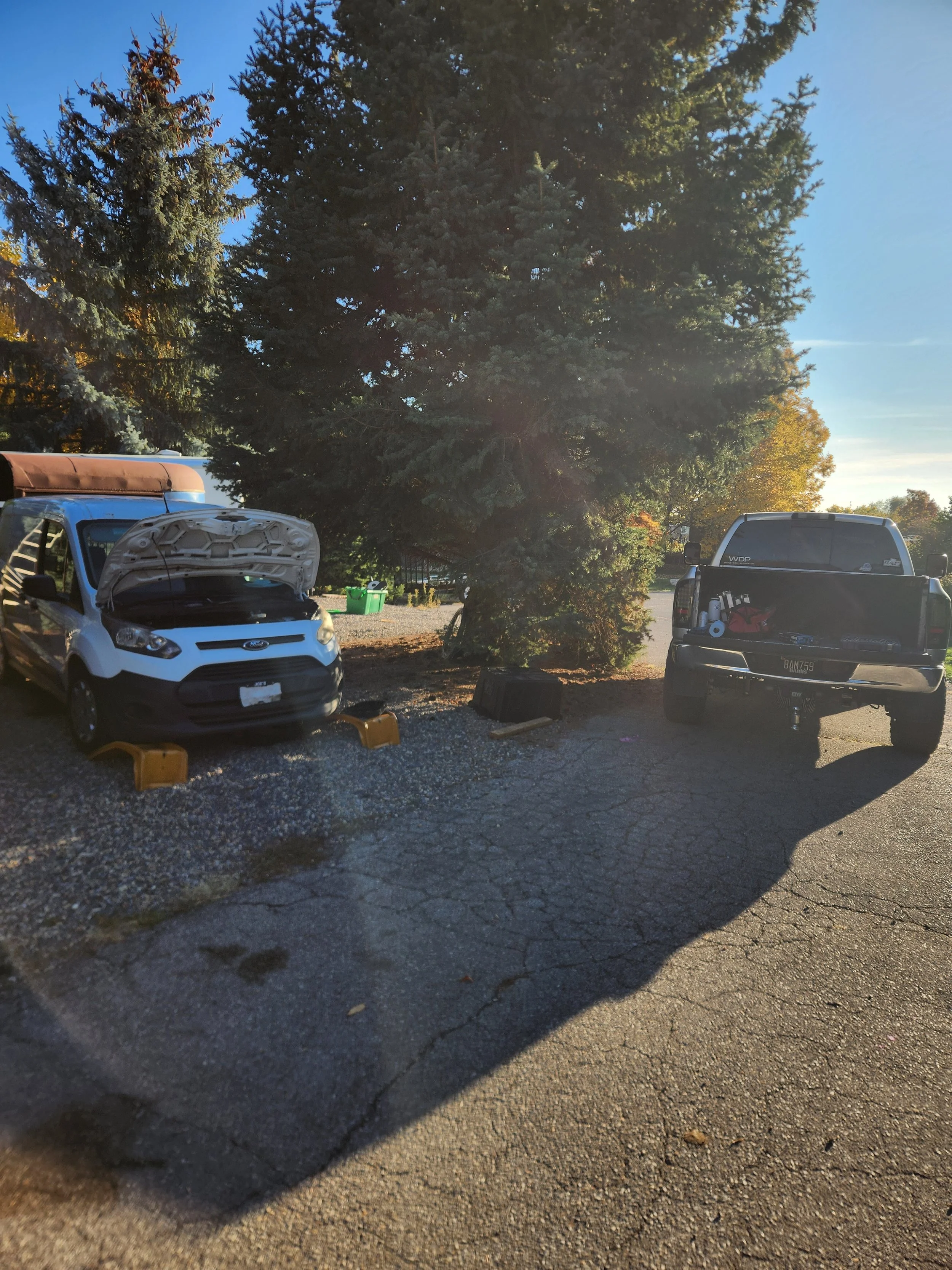 Two vehicles parked near large evergreen trees in a sunlit area. The vehicle on the left has its hood open and is supported with wheel chocks, suggesting it is being worked on. The vehicle on the right is a pickup truck with tools and supplies in the bed.