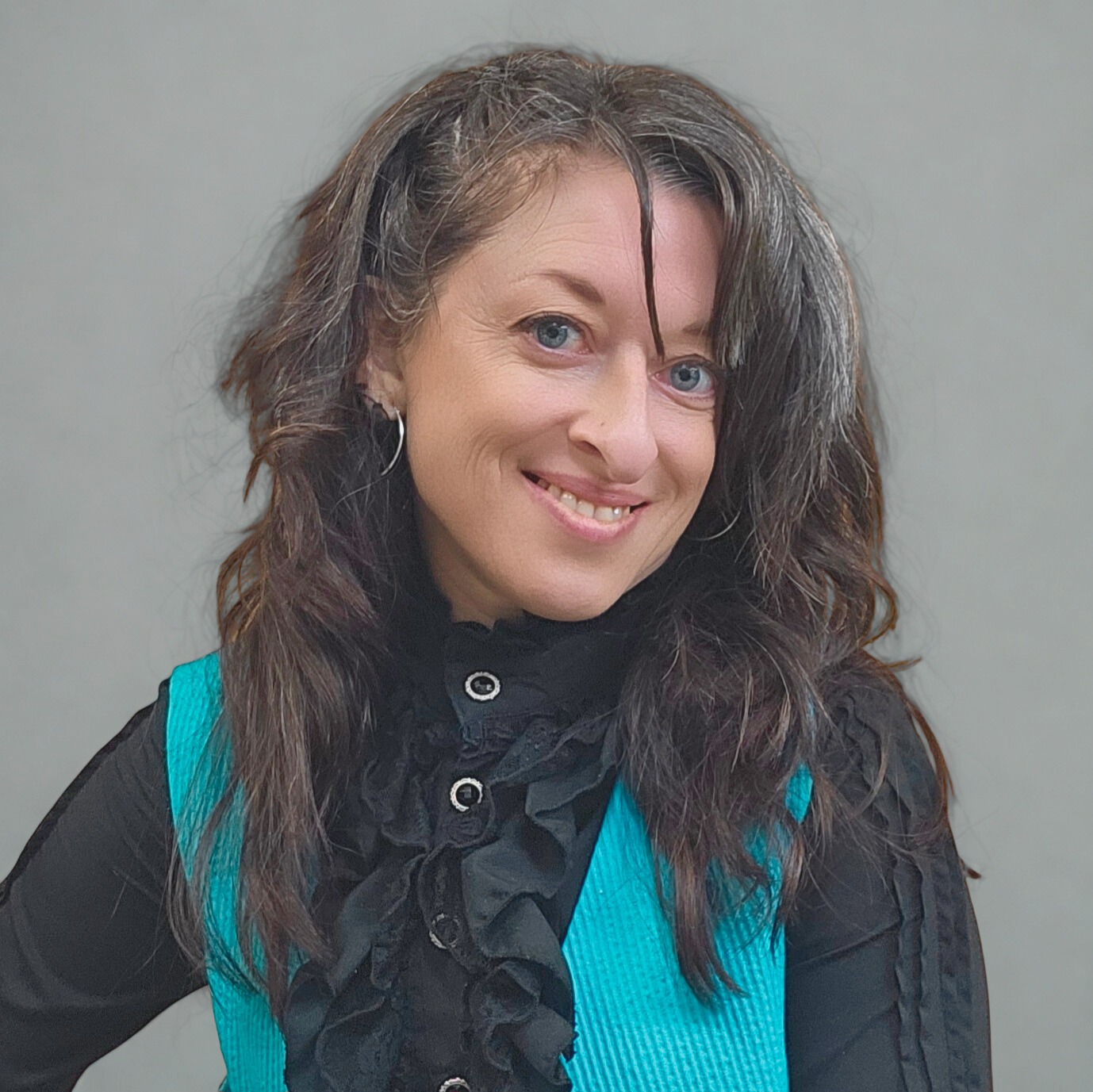 A profile picture of Giselle of Giselle Clothing Design, with shoulder-length wavy brown hair, light skin, wearing a black shirt with ruffled details and a blue vest, standing against a plain grey background.