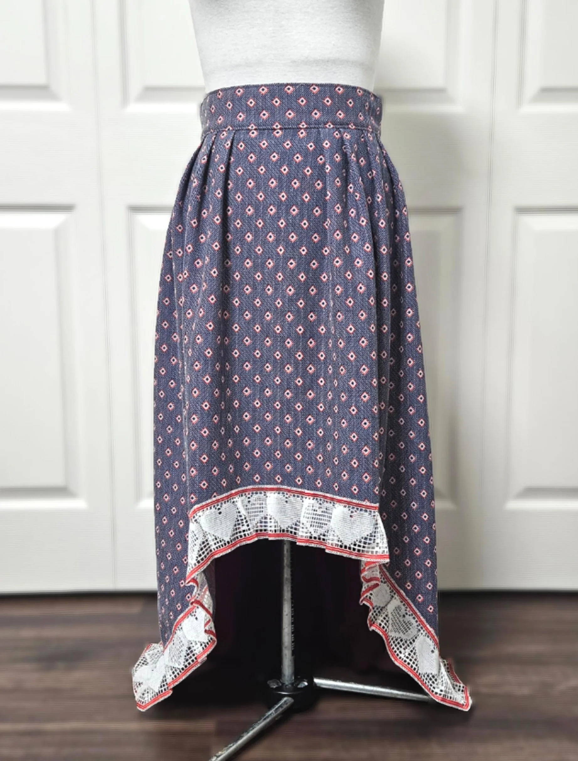 A high-low vintage inspired navy blue skirt with small red diamond patterns and white lace trim at the hem, displayed on a dress form.