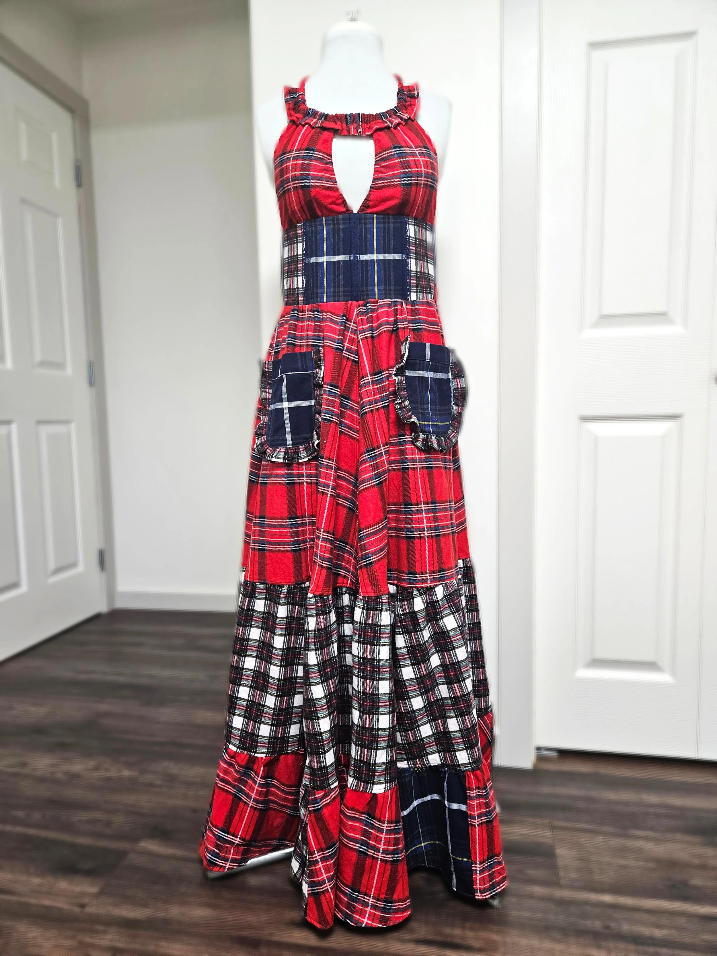 A long, patchwork-style dress with red, navy blue, and white plaid patterns, featuring large pockets and ruffled details, displayed on a white mannequin in a room with wooden floors and white doors.