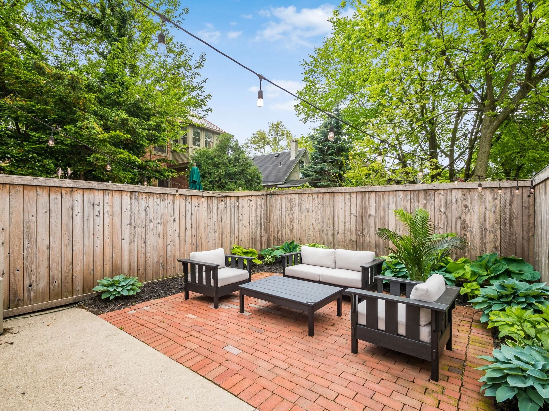 A cozy backyard patio with outdoor seating, including a sofa and armchairs with cushions, surrounded by potted plants and greenery, with a wooden fence and string lights overhead.