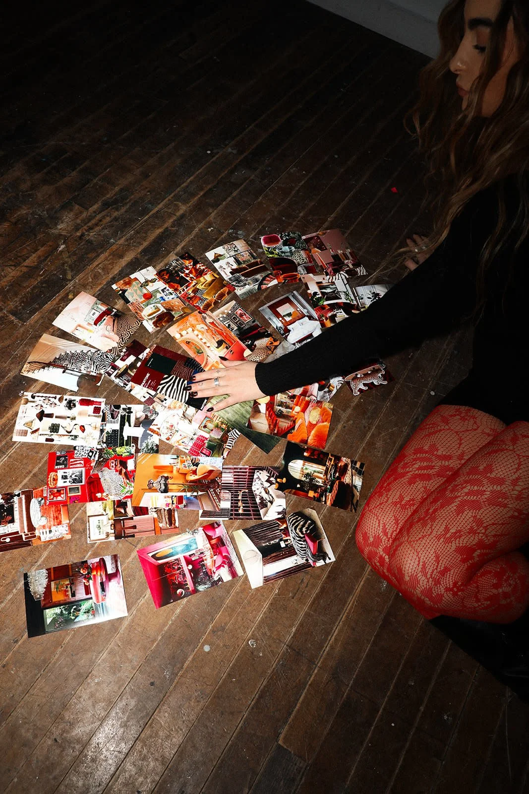 A woman with long hair, wearing a black top and red lace fishnet stockings, seated on a red lacy stool, is arranging a collection of colorful photographs on a wooden floor.